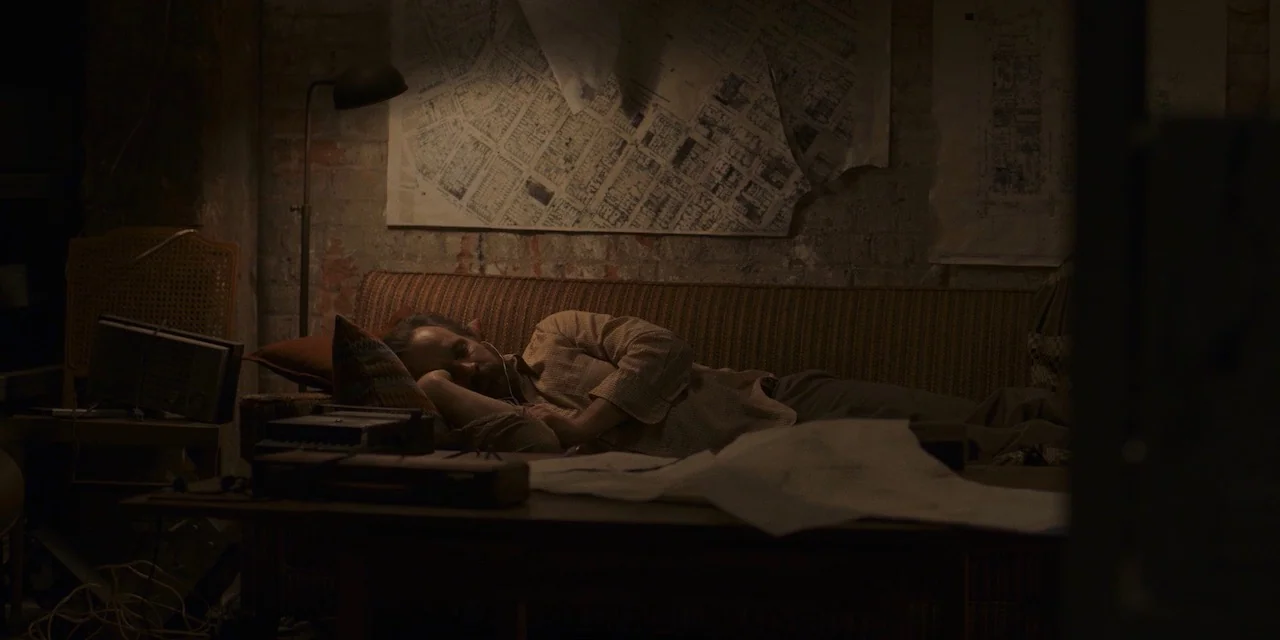 The Sound of Silence movie still: medium — A dimly lit room features a worn sofa against a brick wall with maps. The man lies resting on the couch…