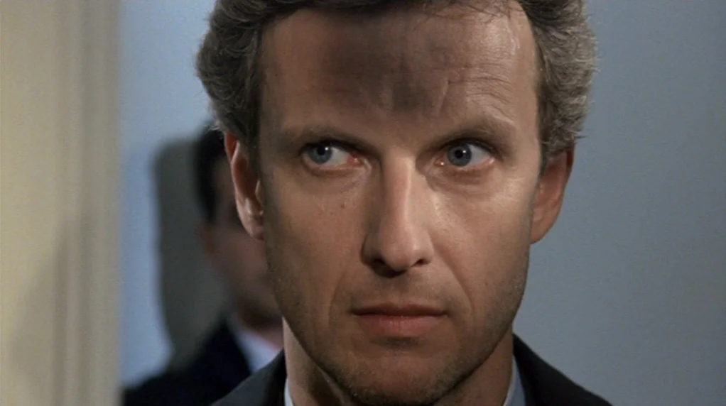Malina (1991) movie still: closeup — An interior office space with a blurred person visible in the background. A man faces forward while looking…