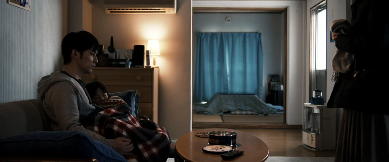 To Hear the Birds Singing (1995) movie still: medium — A quiet Japanese living room contains a sofa and a low coffee table. A man sits with a small…