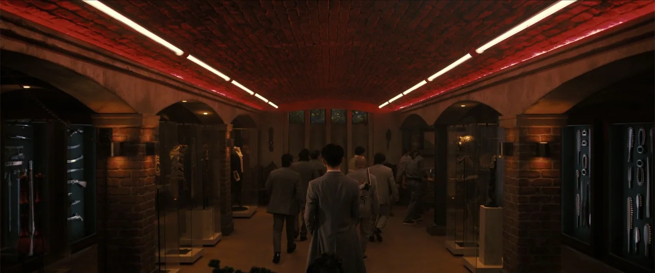 The Wrecking Crew (2026) movie still: wide — A dark museum exhibit hall features brick walls and a vaulted ceiling with red lighting. A group of men…