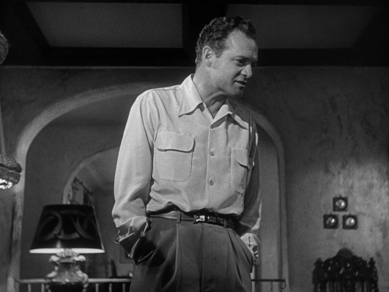 The Prowler (1951) movie still: medium — A dark, furnished domestic interior featuring an arched doorway in the background. A man stands in the…