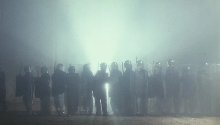 Edward II (1991) movie still: wide — A line of riot police stands in a dark, foggy urban environment at night. They are silhouetted against a…