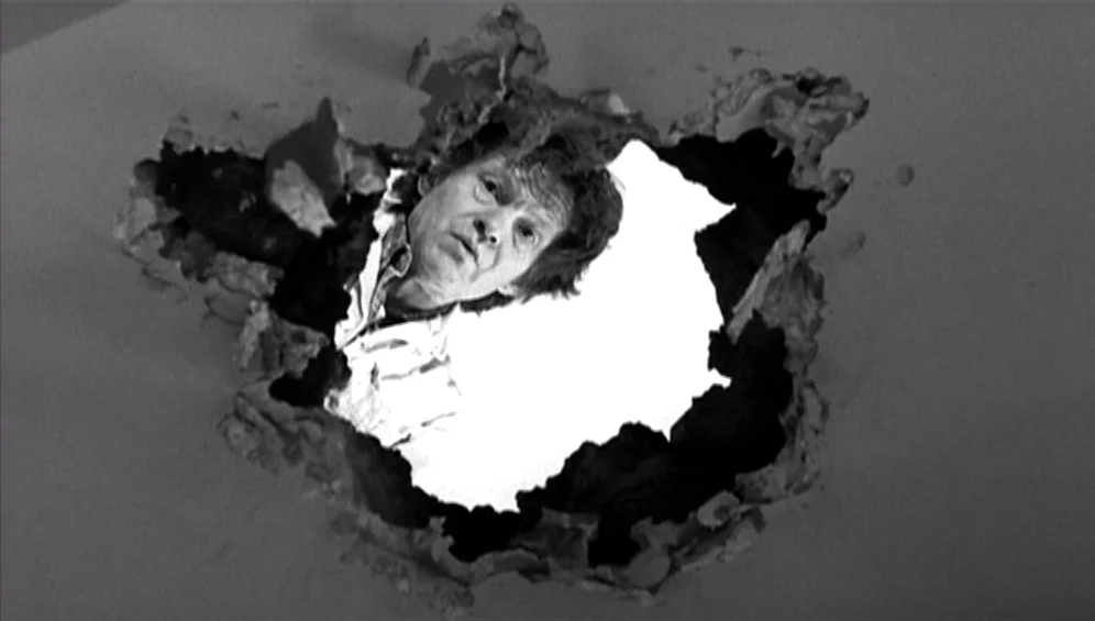 The Tango Lesson (1997) movie still: closeup — A man's face appears through a jagged, irregular hole in a solid material. The hole is surrounded by…