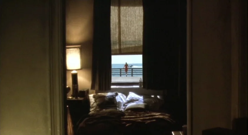 Requiem For A Dream (2000) movie still: wide — A dimly lit room with a messy unmade bed and a table lamp. Through a central window, a woman in a red…