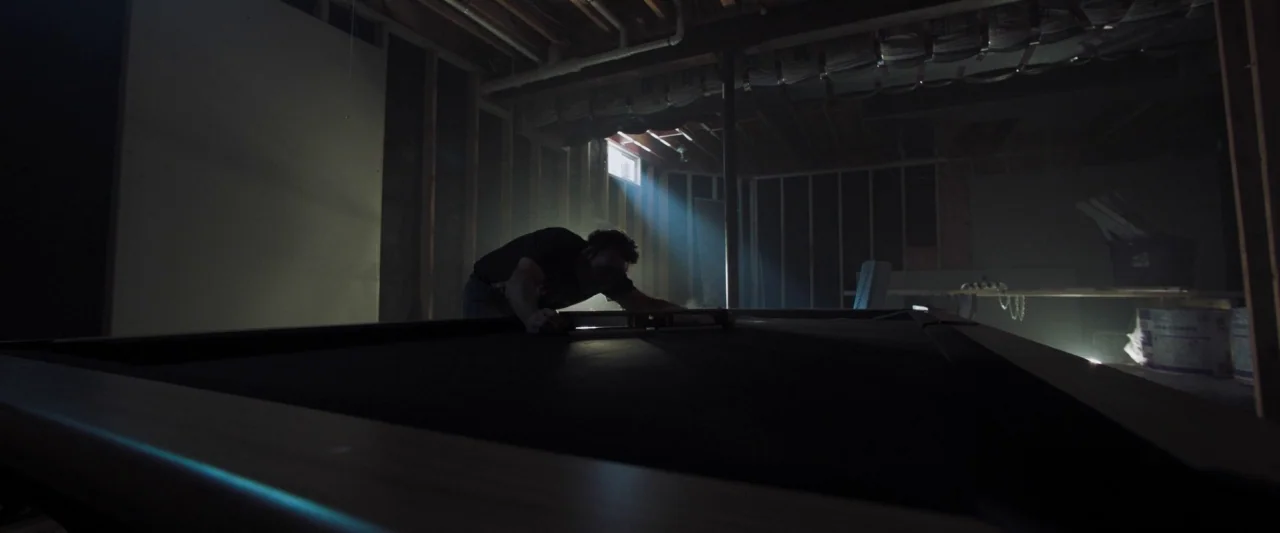 Mom and Dad (2017) movie still: wide — An unfinished basement has exposed wooden wall studs and a low ceiling with joists. A man in a dark shirt…