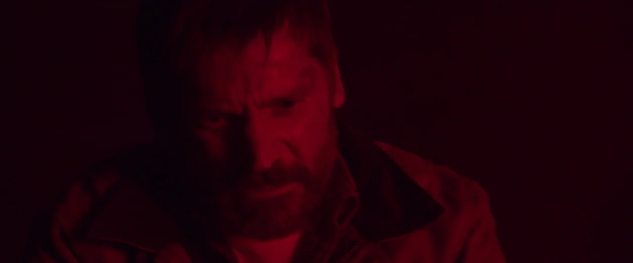 The Silencing (2020) movie still: closeup — A man sits in a dimly lit, nondescript room bathed in dark red light. The man stares downward with a…