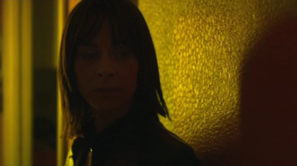 Red Road (2006) movie still: closeup — A dimly lit indoor space with textured wall surfaces colored in vibrant yellow. A woman with dark hair stands…