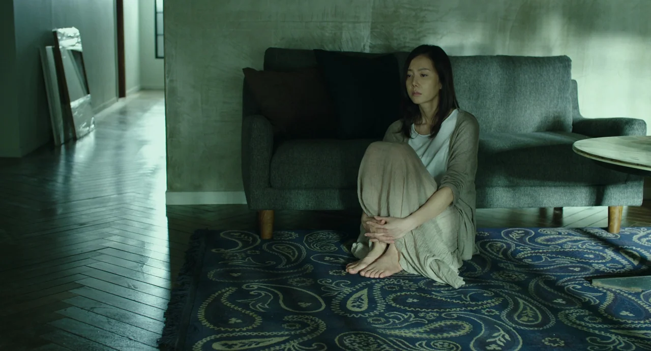 The Mimic (2017) movie still: medium — A modern living room features polished wood floors and a patterned blue rug. A woman sits on the floor…
