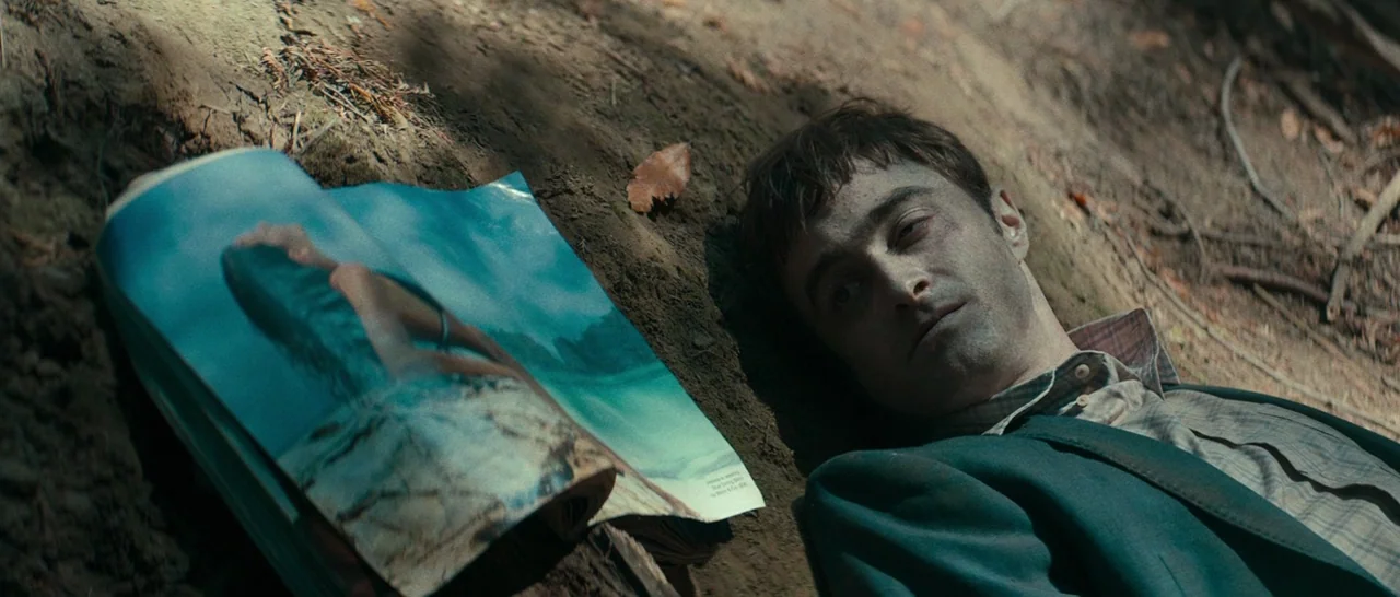 Swiss Army Man (2016) movie still: medium — A man lies flat on a patch of loose brown dirt and pine needles. An open magazine depicting a beach…