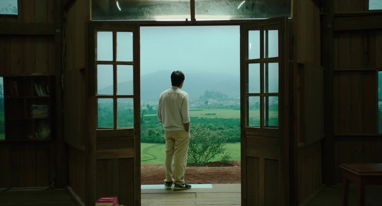Inside The Yellow Cocoon Shell (2023) movie still: fullbody — A wooden cabin interior with exposed beams faces an open landscape of green fields. A…