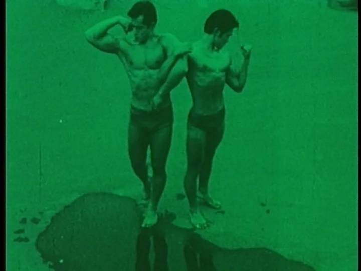 The Cage (1964) movie still: fullbody — Two muscular men in athletic trunks pose on a flat concrete surface. The ground features a dark puddle…