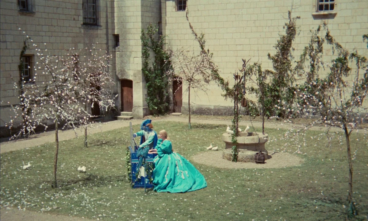 Donkey Skin (1970) movie still: wide — A stone courtyard with climbing ivy walls features a central well and flowering trees. A woman in a large…