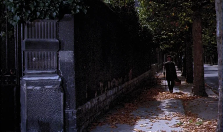 The Thief of Paris (1967) movie still: wide — A sidewalk runs alongside a dark stone wall covered in hanging vines and foliage. A lone man in a top…