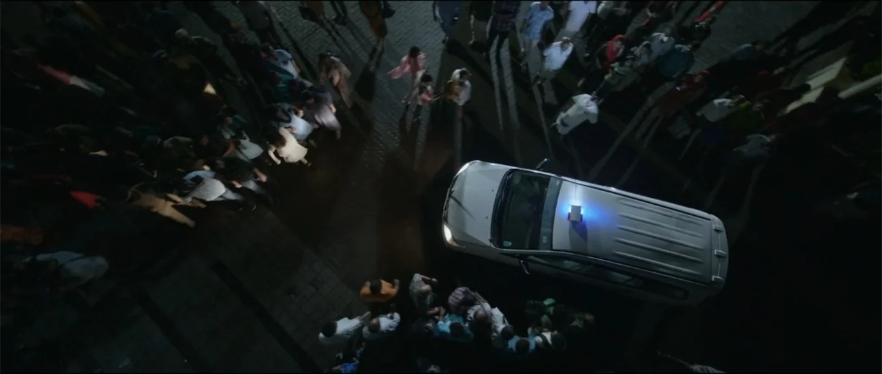 Garudan (2024) movie still: wide — A dark paved outdoor area filled with a large crowd of people at night. A white SUV with flashing blue emergency…