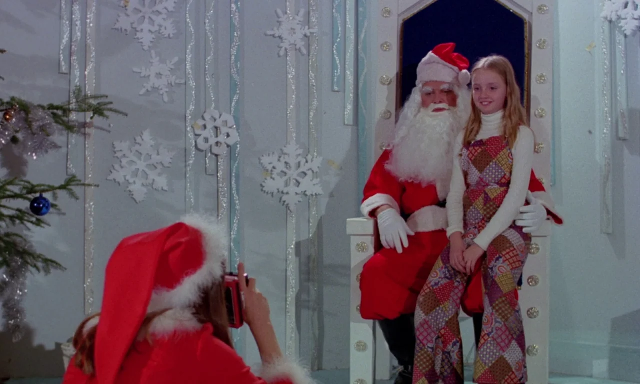 Rabid (1977) movie still: medium — A department store Santa photo setup decorated with white paper snowflake wall ornaments. A young girl takes a…
