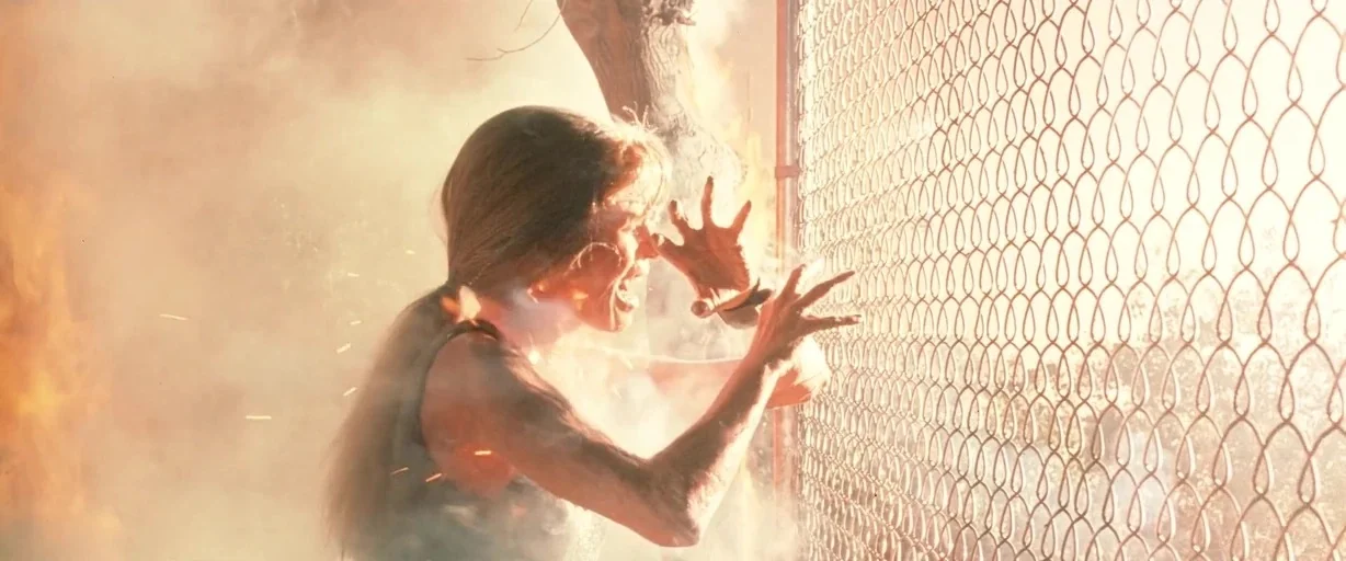 Terminator 2: Judgement Day (1991) movie still: medium — A woman stands near a chain link fence enveloped in thick orange smoke and fire. She…
