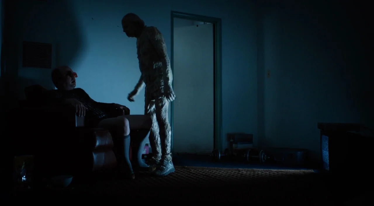 The Greasy Strangler (2016) movie still: wide — A dimly lit room at night features a man sitting on a leather chair. A tall, pale, costumed figure…