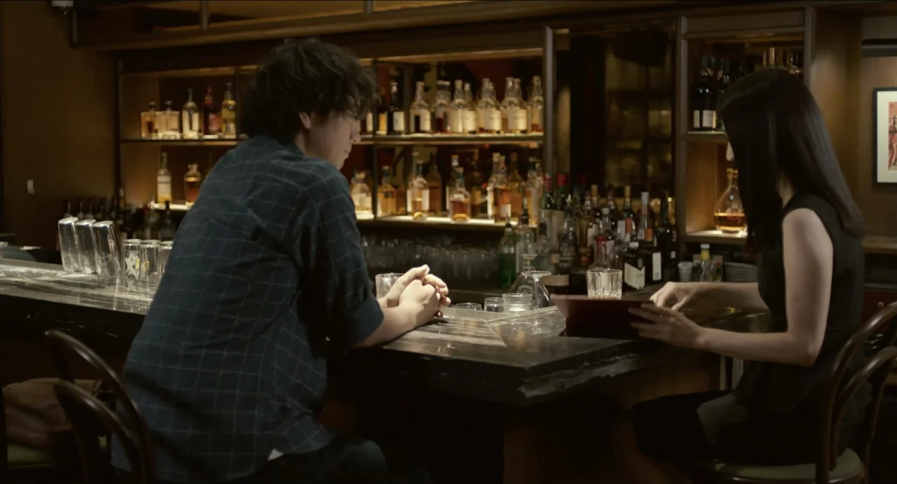 Ramen Shop (2018) movie still: medium — A dimly lit bar with rows of glass liquor bottles on shelves behind. A man and a woman sit at the counter…