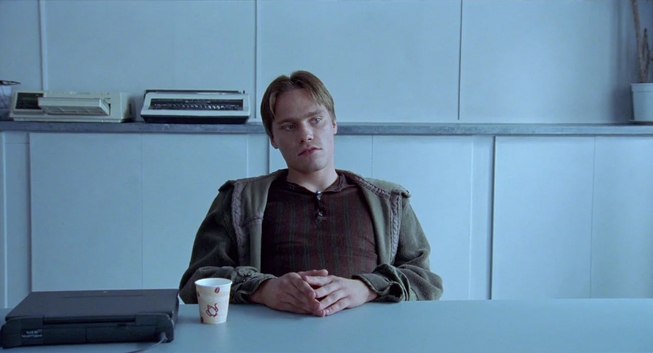 Insomnia (1997) movie still: medium — A man sits at a light blue desk in a sparsely furnished sterile room. He stares forward with clasped hands…