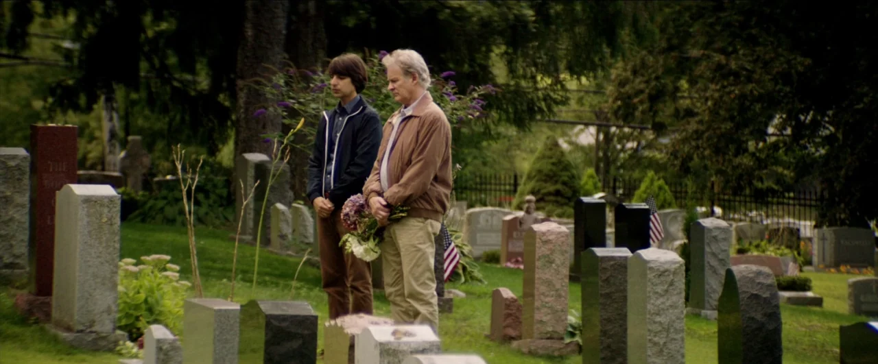 Dean (2016) movie still: fullbody — A green cemetery landscape filled with various granite headstones and lush green trees. Two men stand together…