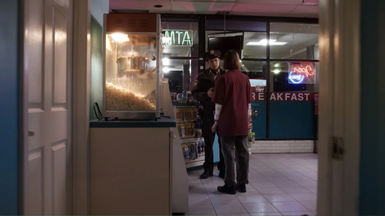 Burn (2019) movie still: medium — A convenience store interior at night features a popcorn machine and glass windows with signs. A police officer in…