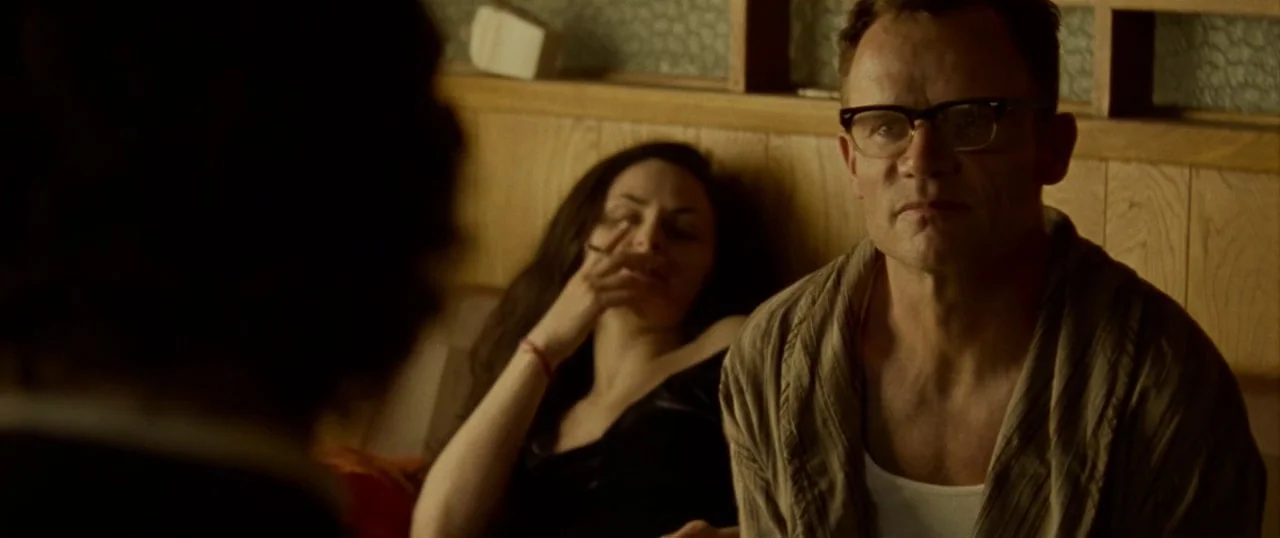Low Down (2014) movie still: medium — A dimly lit space with wood-paneled walls and textured glass room dividers. A man wearing glasses sits in the…