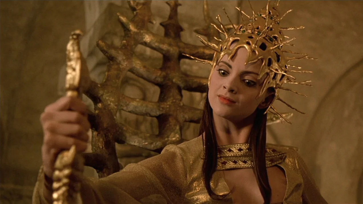 Dagon (2001) movie still: medium — A woman sits in a textured bronze chair within a stone-walled room. She holds a golden ornate scepter while…