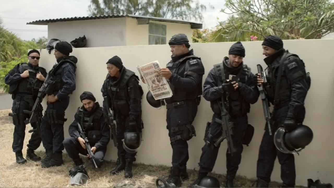 Unknown #1 movie still: fullbody — A group of armed tactical officers waits against a long concrete perimeter wall outdoors. Several officers are…