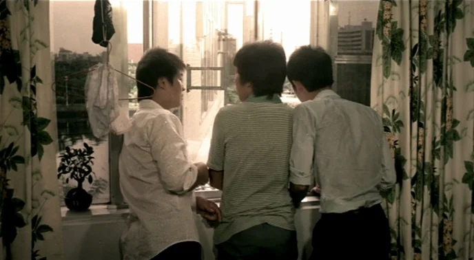 The Boys From Fengkuei (1983) movie still: medium — A residential room features floral curtains framing a window looking out onto a city view. Three…