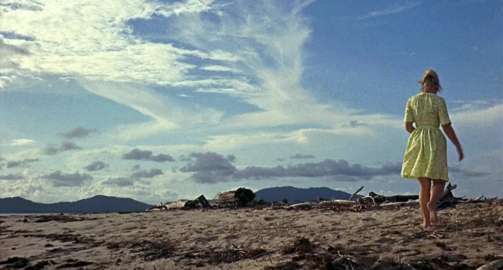 Age of Consent (1969) movie still: fullbody — A wide sandy beach stretches toward distant mountains under a blue sky with clouds. A woman in a light…