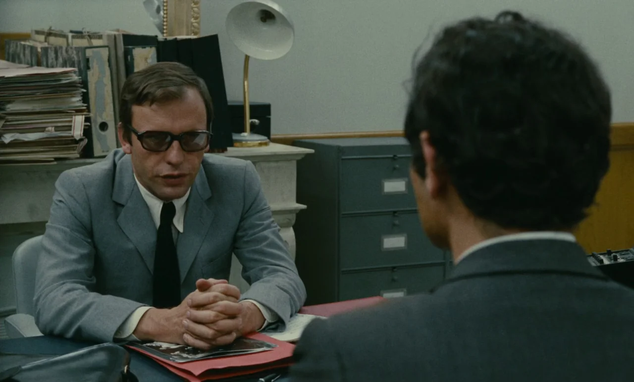 Z (1969) movie still: medium — An office features metal filing cabinets and stacks of paper on a desk. A man in a suit sits behind the desk talking…