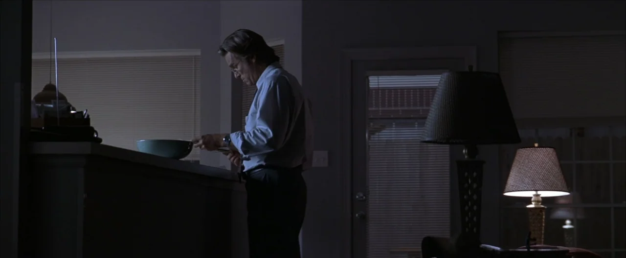 Arlington Road (1999) movie still: medium — A dark, sparsely furnished interior features a man standing near a kitchen counter. The space is…