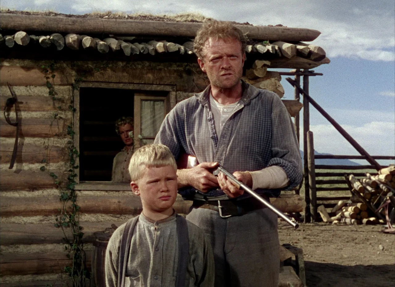Shane (1953) movie still: medium — A rustic log cabin with a grass-covered roof sits under a cloudy sky. An adult male with a rifle stands in front…