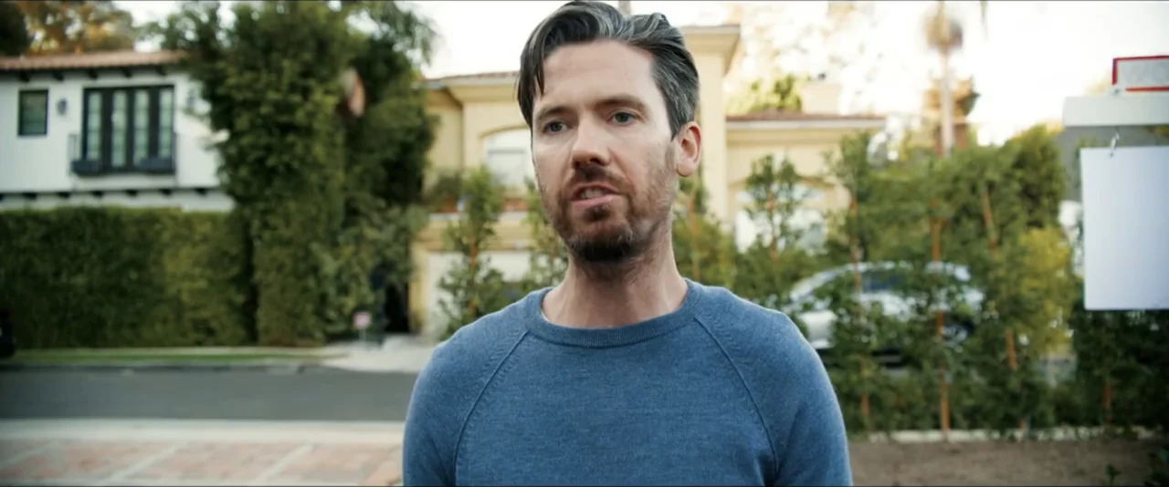 Gatlopp: Hell of a Game (2022) movie still: medium — A suburban street in front of a tan house with tall hedges and trees. A man in a blue sweater…