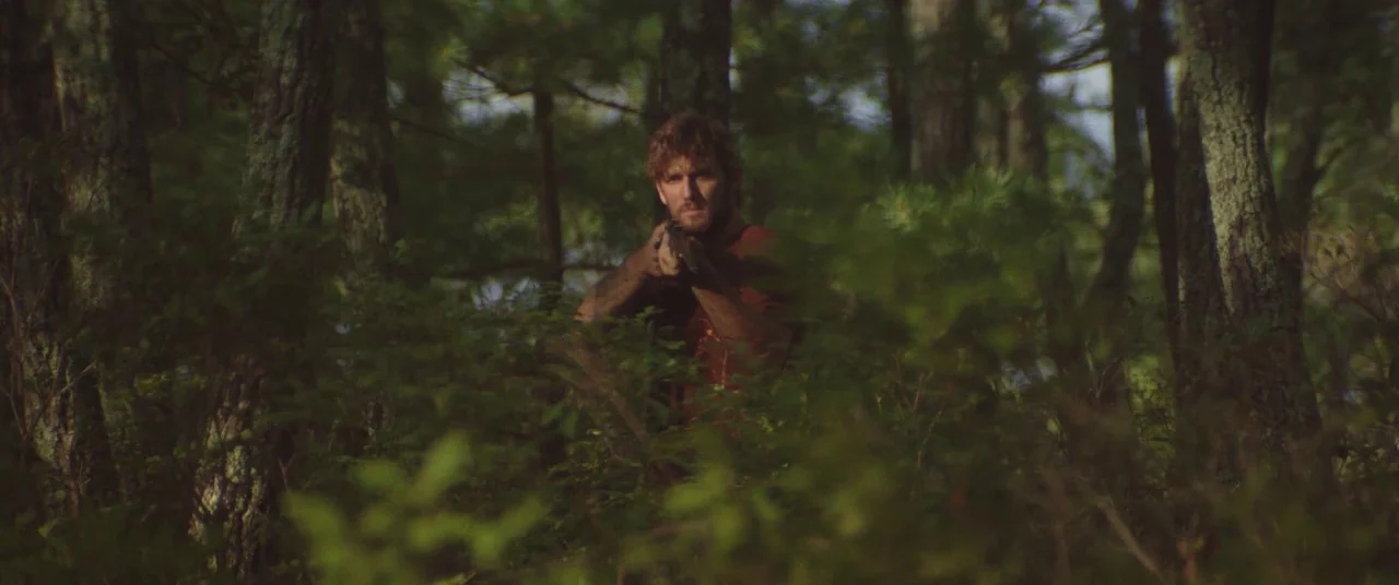 The Strange Ones (2017) movie still: medium — A dense woodland with thick green foliage and vertical tree trunks in the background. A man stands…