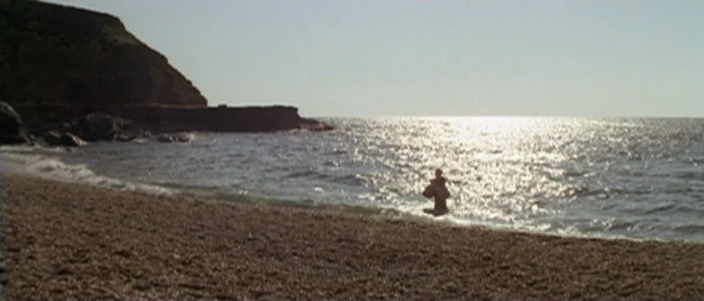 Boom! (1968) movie still: wide — A desolate pebble beach stretches along a calm sea under a bright, cloudless sky. A woman stands waist-deep in the…