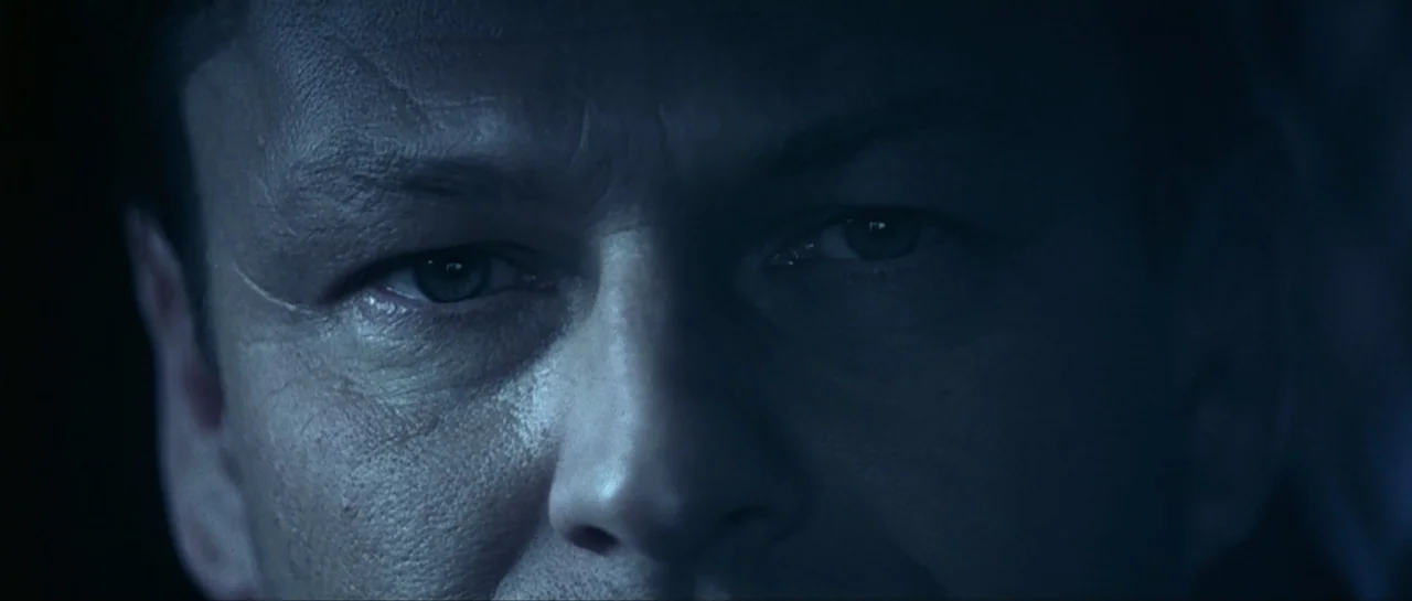 Don't Say a Word (2001) movie still: closeup — A man's face fills the frame in a dark, dimly lit environment. He looks forward with a neutral…