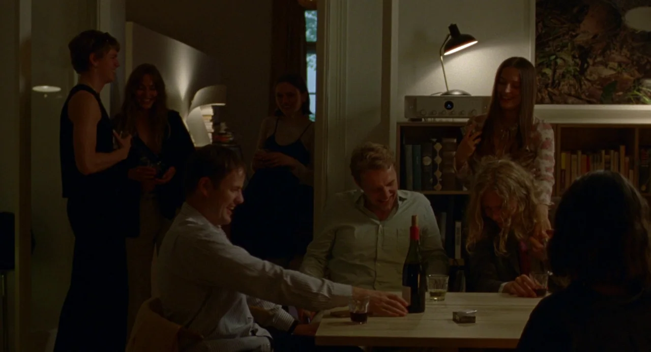Sick of Myself (2022) movie still: medium — A dimly lit room holds a group of people gathered around a wooden table. The subjects are engaged in…