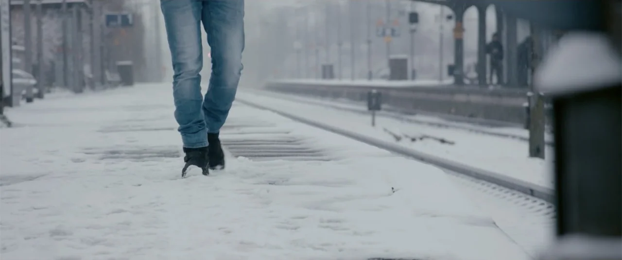Simple as Water (2021) movie still: medium — A snowy train track platform in an industrial setting with blurred structures in distance. A person in…