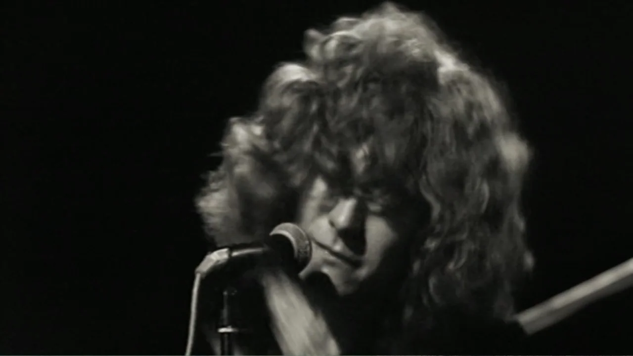 Becoming Led Zeppelin (2025) movie still: closeup — A dark concert stage interior features a performer in a close shot. The man sings with his eyes…
