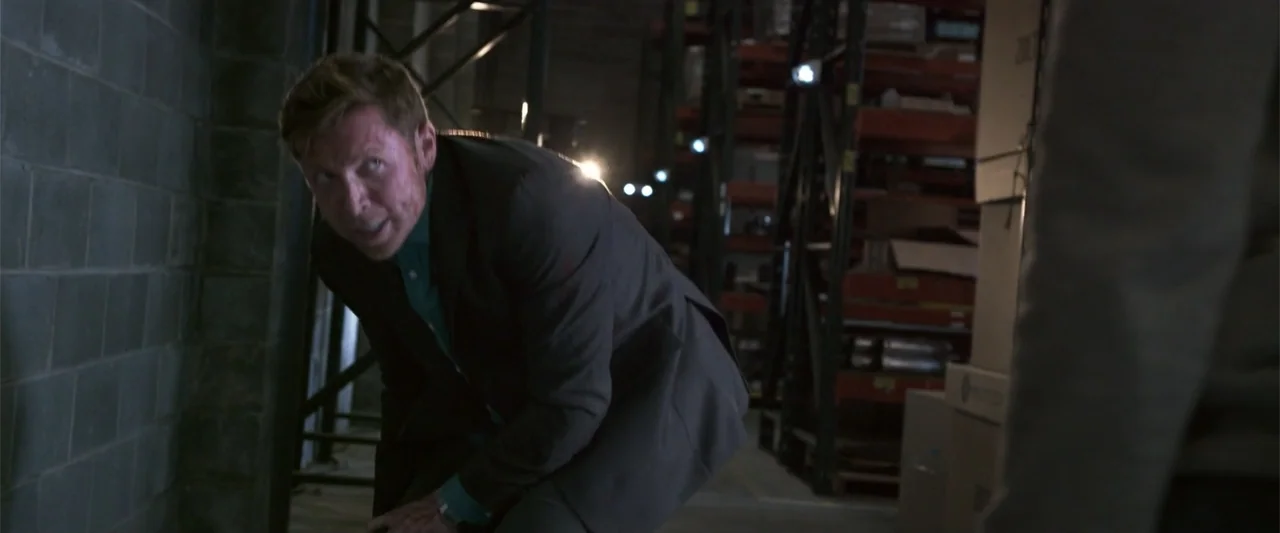 Office Uprising (2018) movie still: medium — A dark warehouse interior contains stacked cardboard boxes and metal shelving units in shadow. A man in…