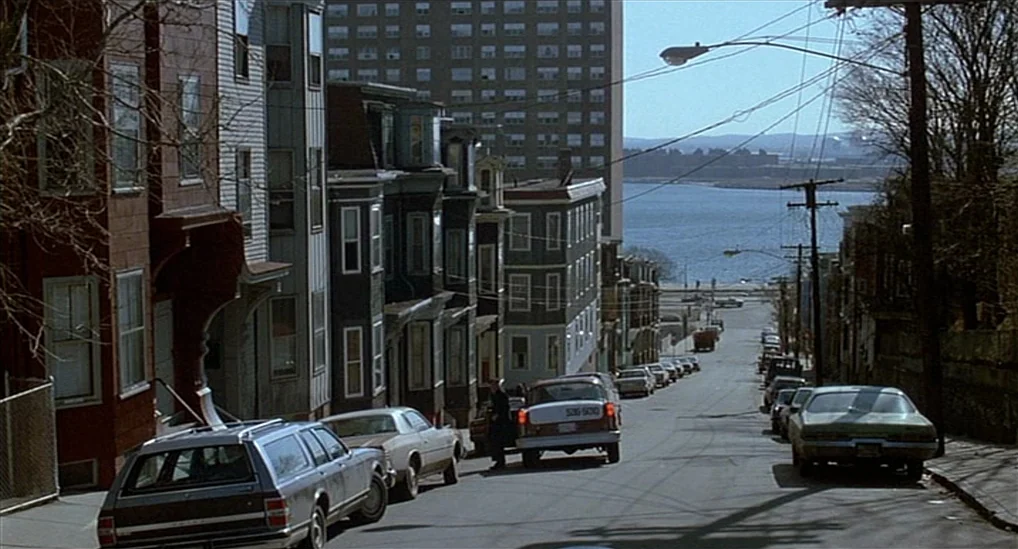 The Verdict (1982) movie still: establishing — A steeply sloped residential street is lined with multi-story wooden apartment buildings and parked…
