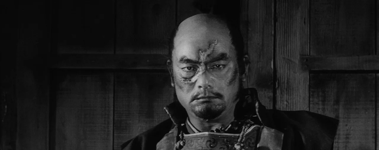 The Hidden Fortress (1958) movie still: closeup — A man stands in front of a dark, vertical wooden wall paneling. He stares forward with a visible…