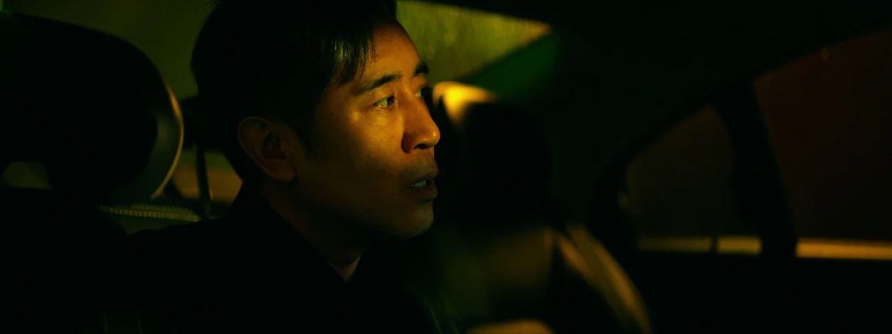 Under The Light (2023) movie still: closeup — The interior of a dark car at night with a man sitting in the back. Yellow light streams through the…