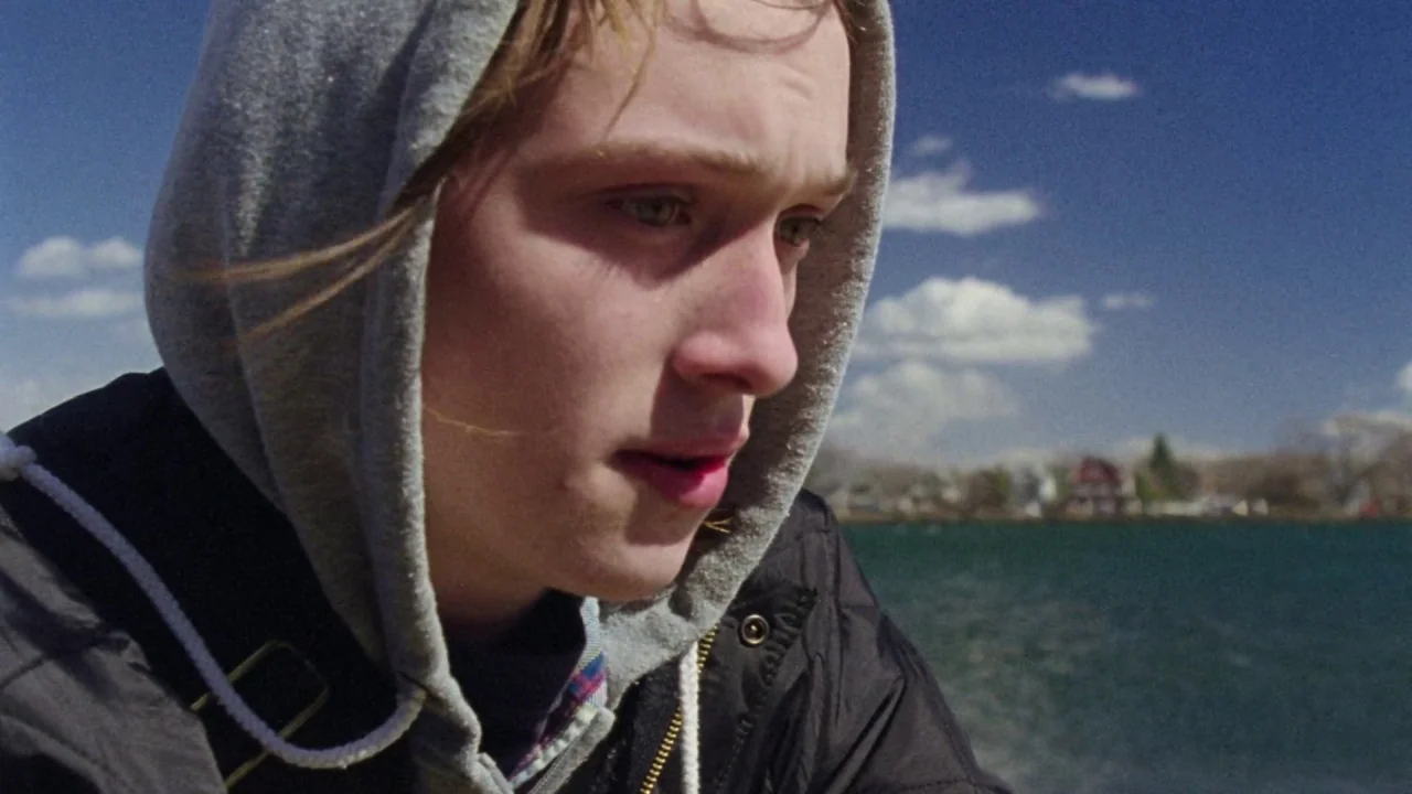 I Am Not a Serial Killer (2016) movie still: closeup — A person stands beside a large lake under a clear, bright blue sky. The subject wears a…