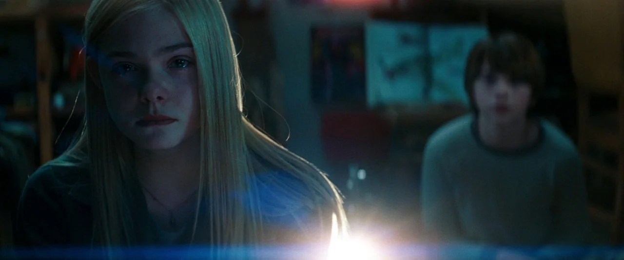 Super 8 (2011) movie still: medium — A dimly lit room at night with posters visible on the back wall. Two teenagers sit near a light source that…