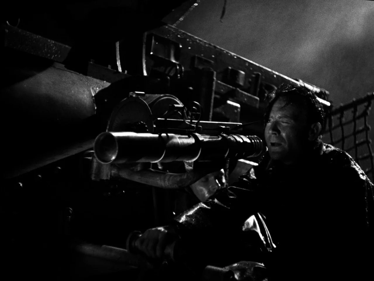 In Which We Serve (1942) movie still: medium — A dark, wet exterior deck of a warship at night with heavy machinery present. A man in a drenched…