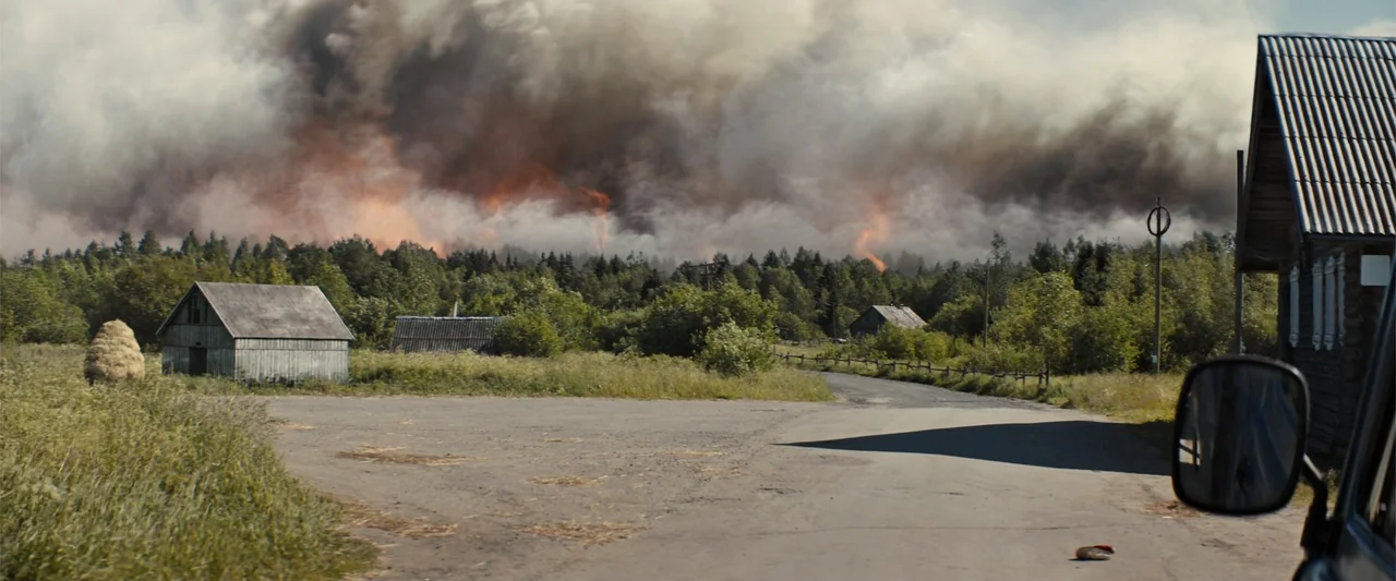 In the Line of Duty: Smoke Jumpers (1996) movie still: wide — A rural road winds through a field with weathered wooden barns and dense forest. A…