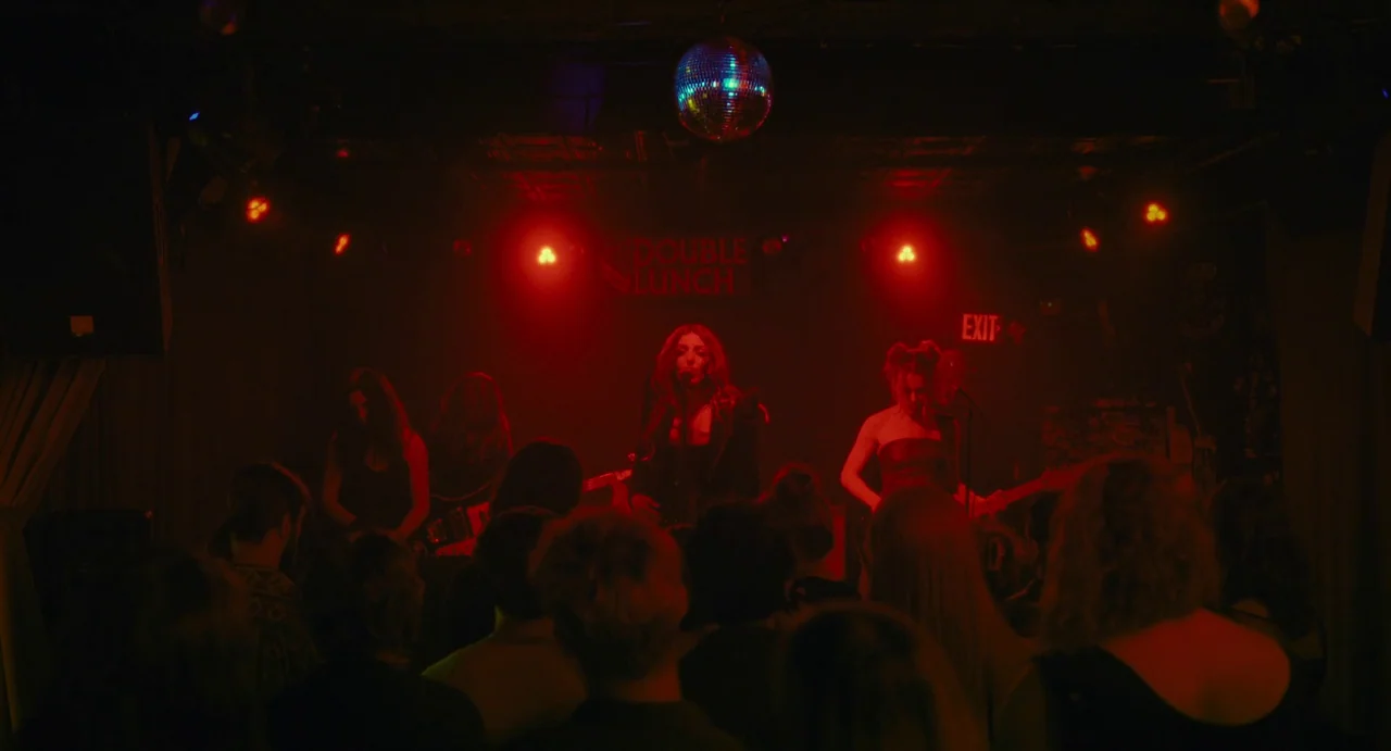 I Saw The TV Glow (2024) movie still: medium — A dark concert club interior featuring a stage with a disco ball overhead. A band performs for a…