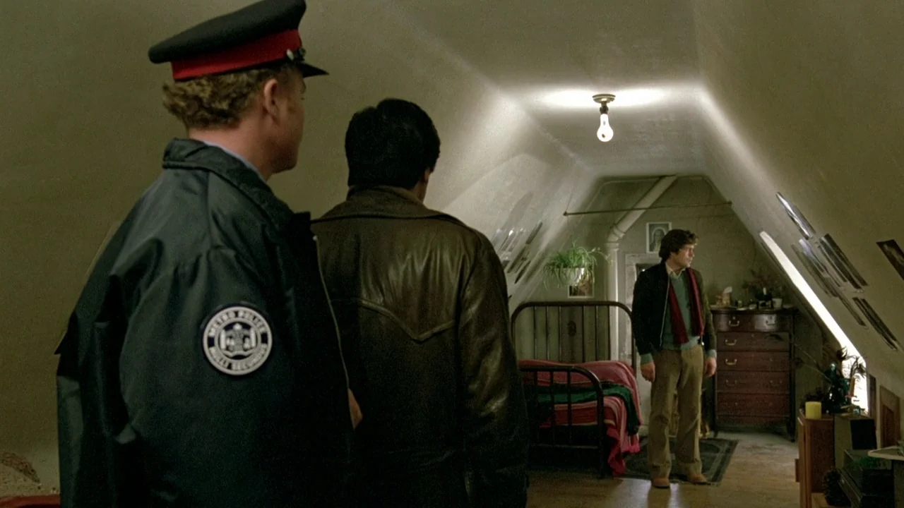 The Brood (1979) movie still: medium — An attic space features slanted walls and a single bulb illuminating a bed. Two men stand looking toward a…